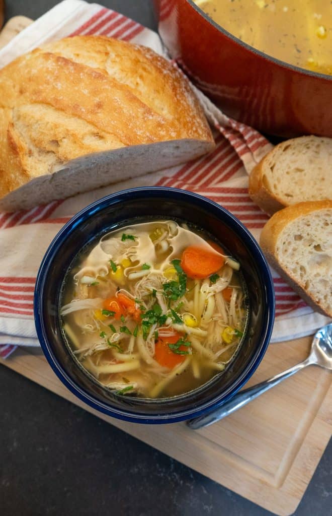 blue bowl full of chicken noodle soup wit carrots and celery visible, a half loaf of bread is beside the bowl and the pot of soup is in the upper right hand corner
