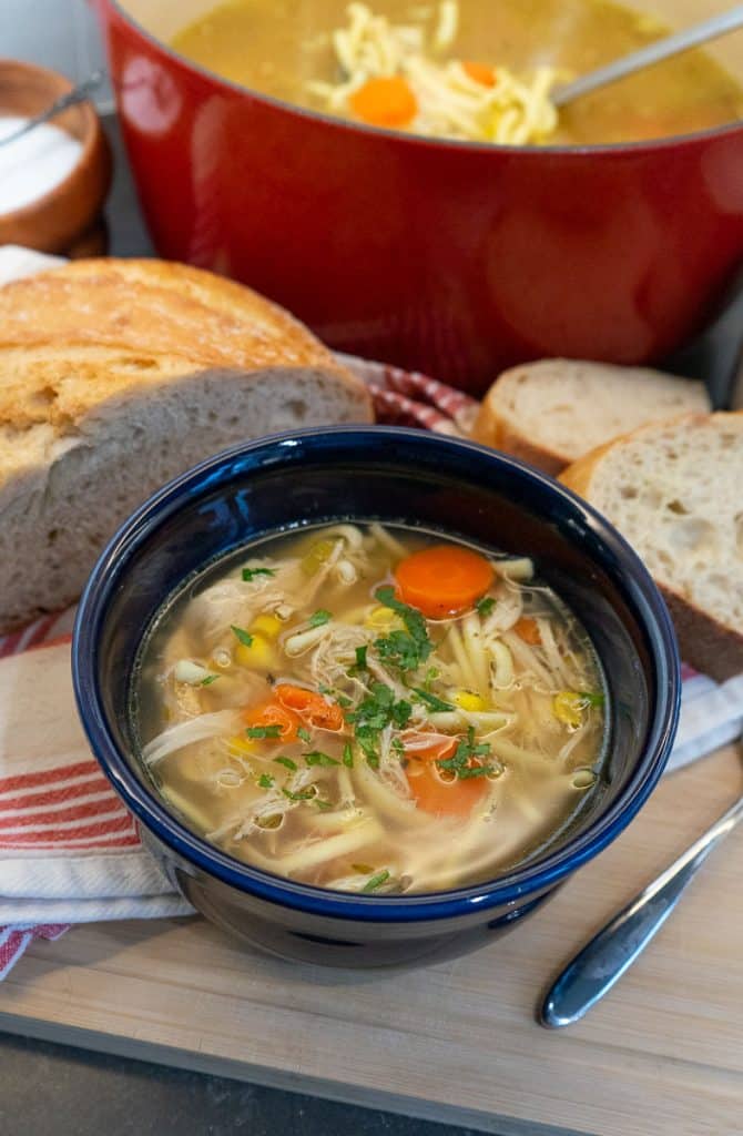blue bowl full of chicken noodle soup wit carrots and celery visible, a half loaf of bread is beside the bowl and the pot of soup is in the upper right hand corner