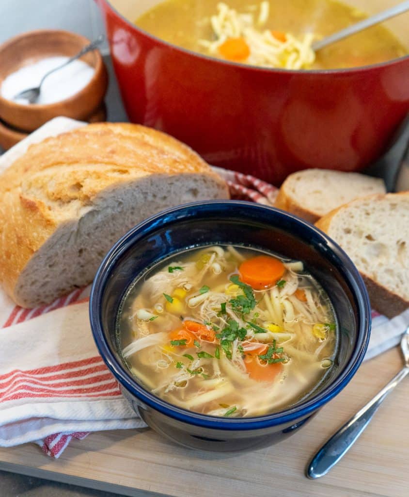 chicken noodle soup in a blue bowl with a loaf of bread and a red dutch oven of soup behind it