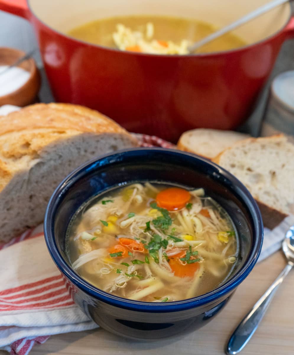blue bowl full of chicken noodle soup wit carrots and celery visible, a half loaf of bread is beside the bowl and the pot of soup is in the upper right hand corner