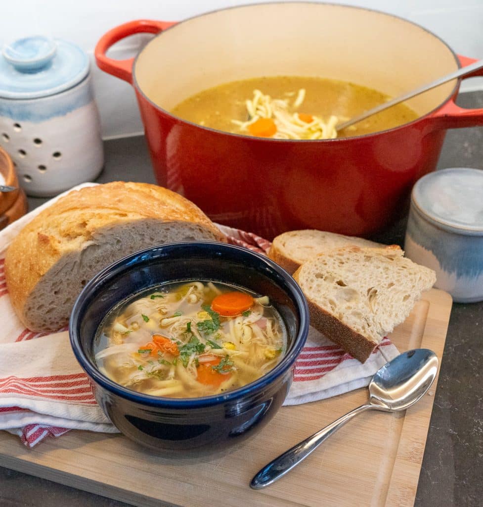 blue bowl full of chicken noodle soup wit carrots and celery visible, a half loaf of bread is beside the bowl and the pot of soup is in the upper right hand corner
