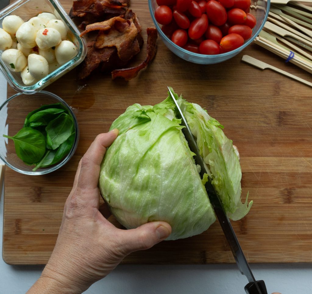 slicing the bottom of a head of iceberg lettuce, on a wood cutting board small glass dishes of mozzarella, bacon, basil and cherry tomatoes above lettuce