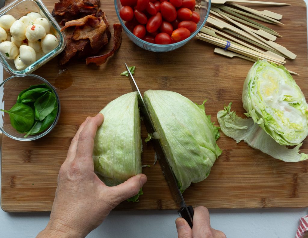 cutting head of iceberg lettuce half on a wood cutting board with glass bowls of cherry tomatoes, bacon, mozzarella and basil above it