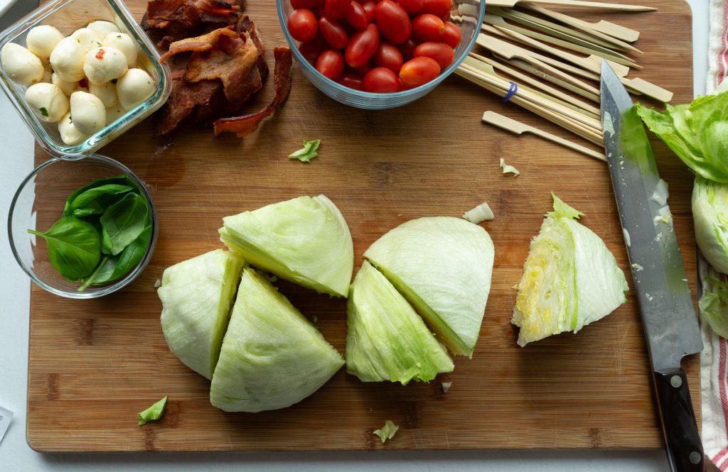 6 wedges of iceberg lettuce on a wood cutting board with glass bowls of cherry tomatoes, bacon, basil and mozzarella above