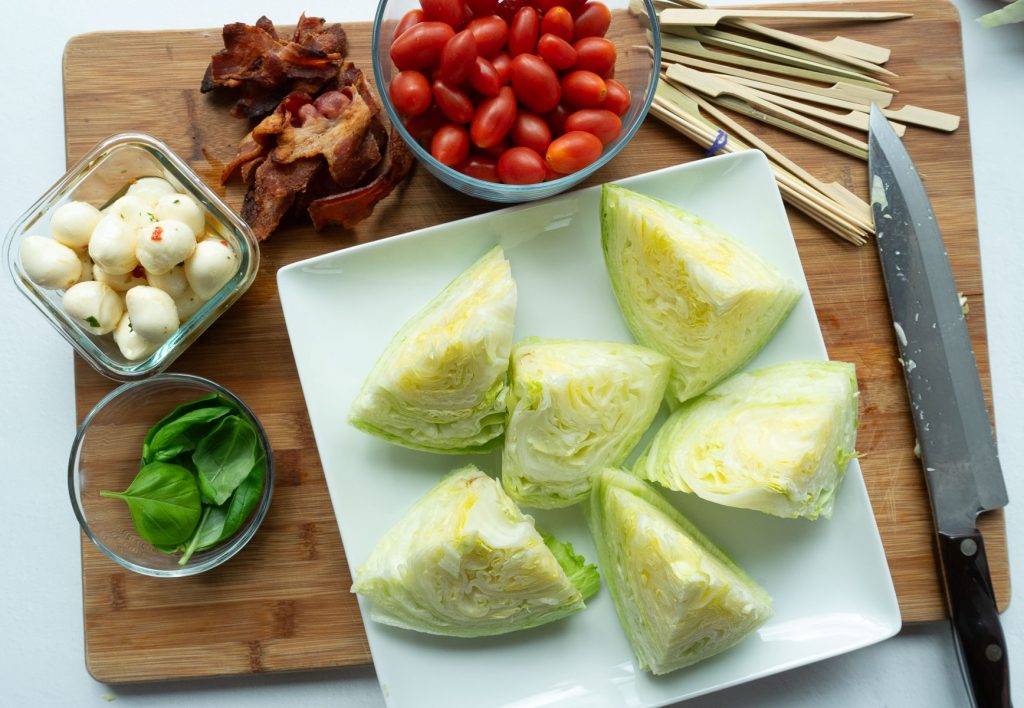 assembling mini wedge salad skewers. lettuce wedges on white platter on top of a wood cutting board with ingredients surrounding the white platter