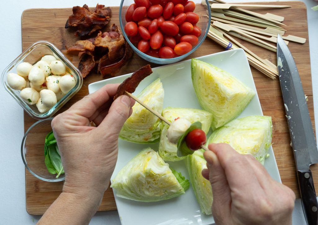 skewering tomato, mozzarella, basil, and bacon above a white platter of iceberg lettuce wedges