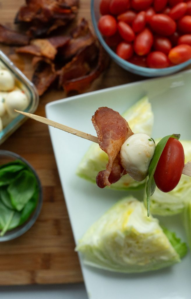 tomato, basil, mozzarella ball, bacon on a wood skewer above white platter of iceberg lettuce wedges