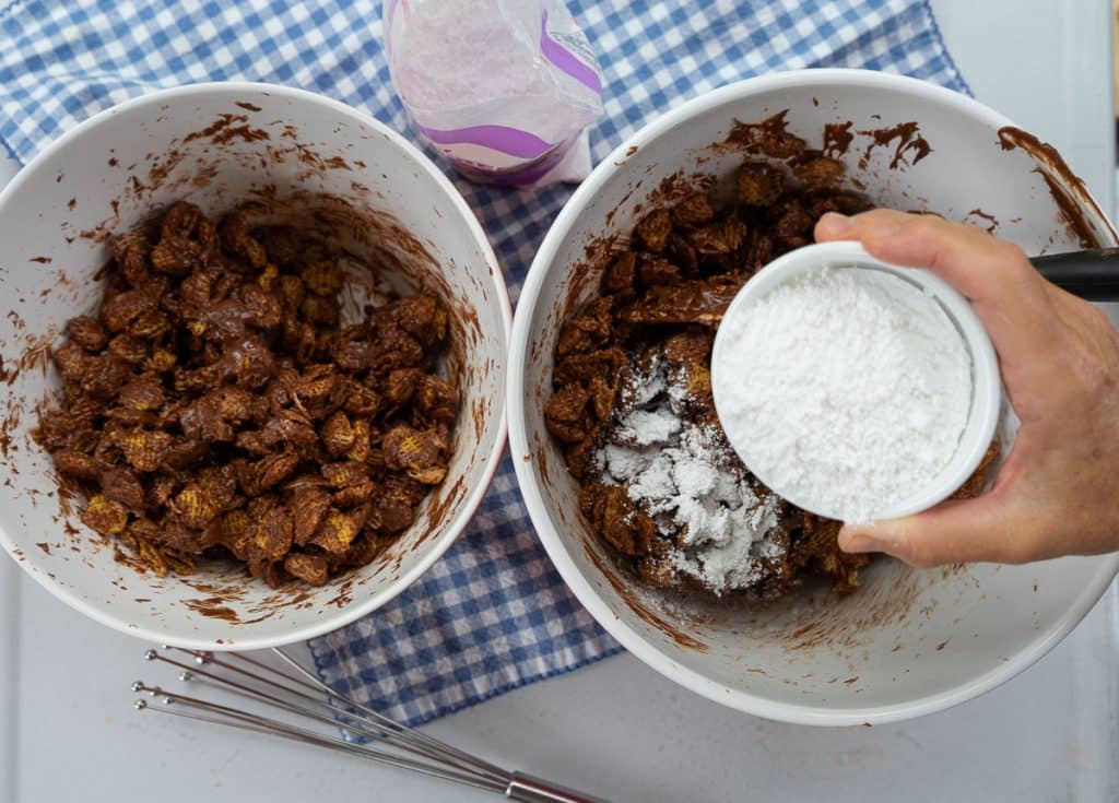 two bowls of crispex cereal mixed with chocolate mixture with a hand coming in from the right starting to sprinkle powdered sugar over the cereal