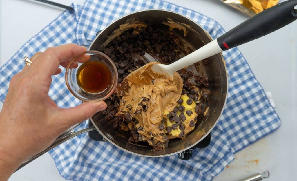 chocolate chips, butter, and peanut butter melting in a saucepan with a hand coming in from the left adding vanilla. there is a spatula in pan