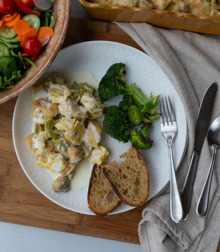 plated chicken tortellini bake on a white plate with broccoli and garlic toast, a dish of salad is in the upper left corner and the dish of tortellini is in the upper right corner of the photo