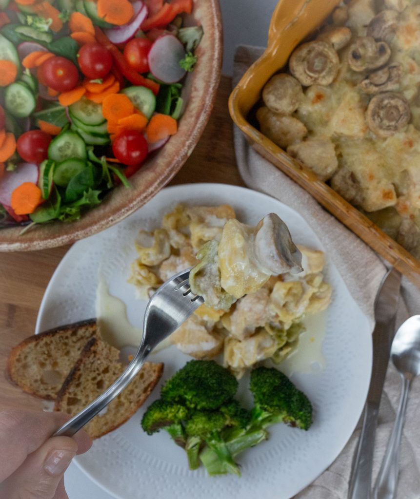 plated chicken tortellini bake on a white plate with broccoli and garlic toast, a dish of salad is in the upper left corner and the dish of tortellini is in the upper right corner of the photo. A fork is in center of photo over plate with tortellini chicken on it