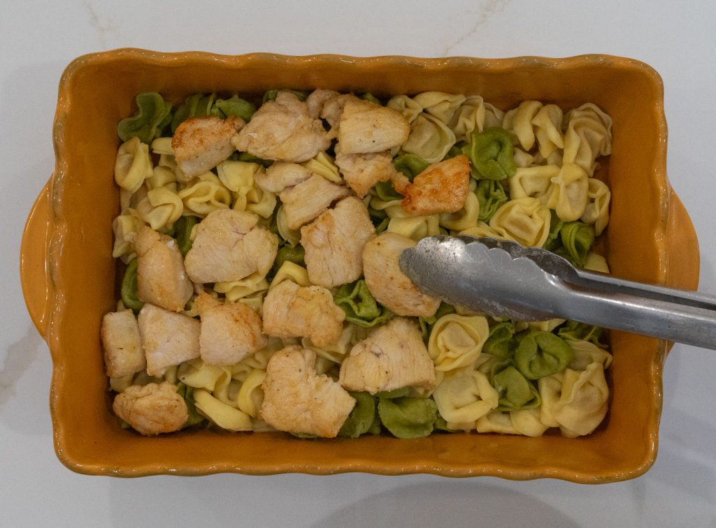 tongs placing chicken on top of tortellini in a gold baking dish