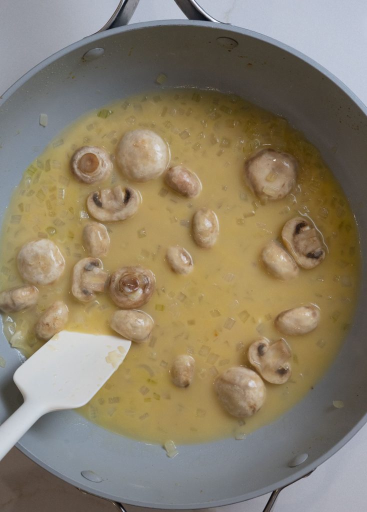 mushrooms and onions cooking in chicken broth in large skillet, a white spatula is in the lower left of the photo