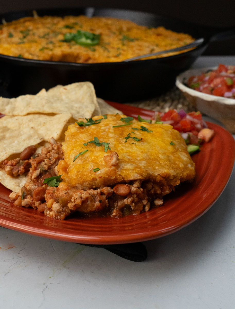 serving of tamale pie on a rust colored fiesta ware plate with tortilla chips on the side pan of tamele pie is in the background