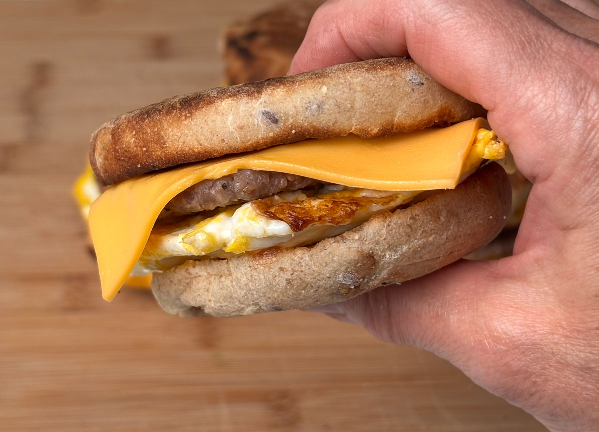 hand holding a sausage and egg breakfast sandwich on an english muffin wood cutting board in the background