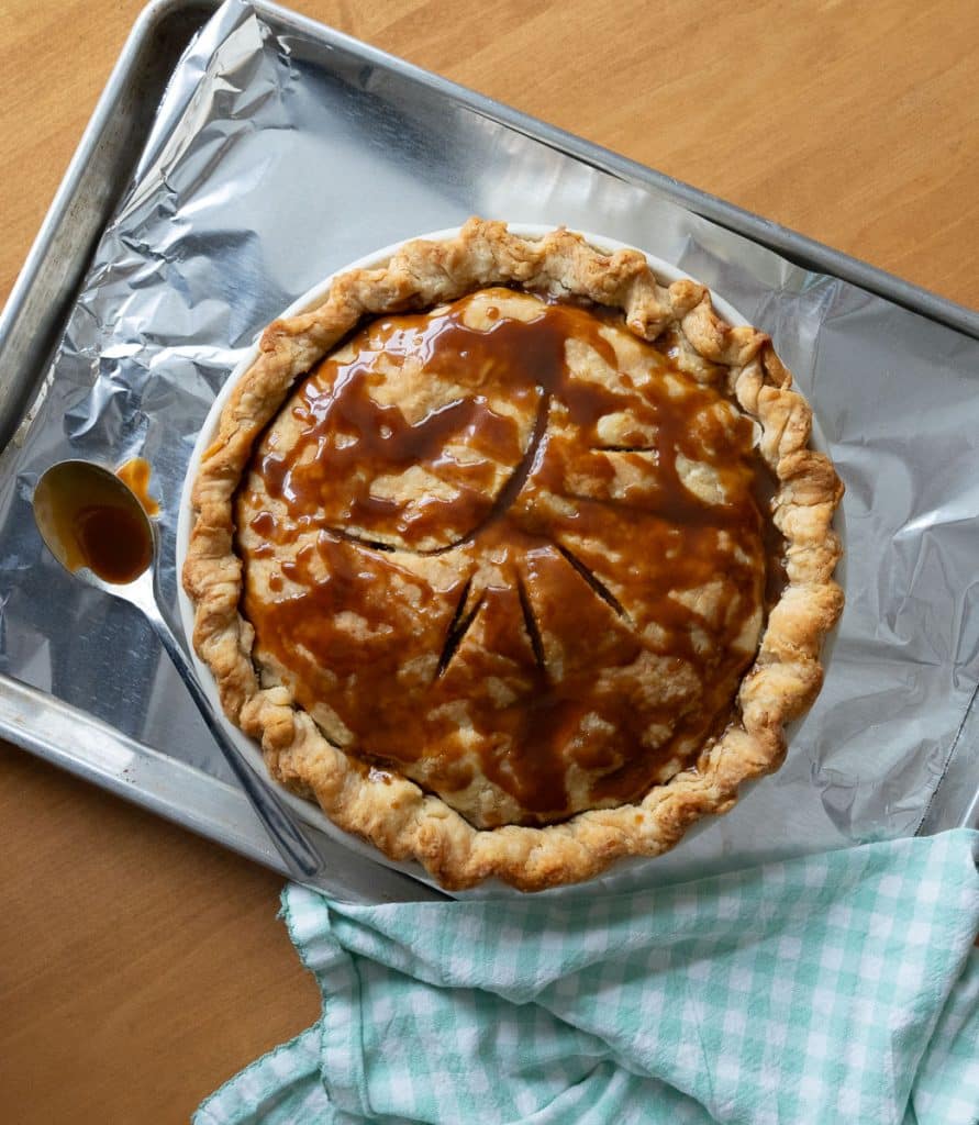 whole caramel apple crunch pie sitting on a baking sheet with a spoon of caramel laying to the lower left of pie on the pan a gingham napkin in the bottom right corner