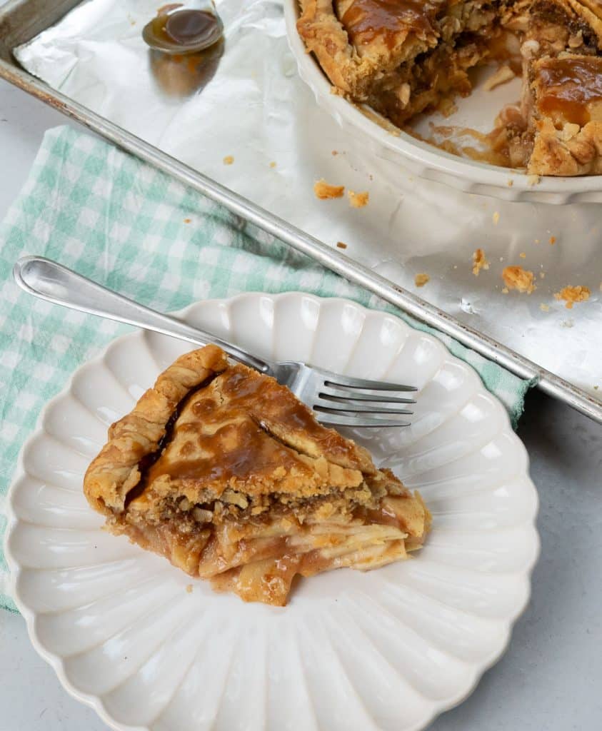 Slice of caramel apple crunch pie on a white plate with a light teal and white gingham napkin under the plate, fork laying on plate, whole pie is in the background on a baking sheet