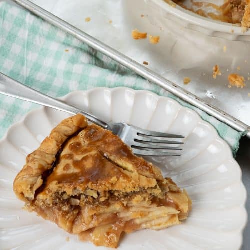 Slice of caramel apple crunch pie on a white plate with a light teal and white gingham napkin under the plate, fork laying on plate, whole pie is in the background on a baking sheet