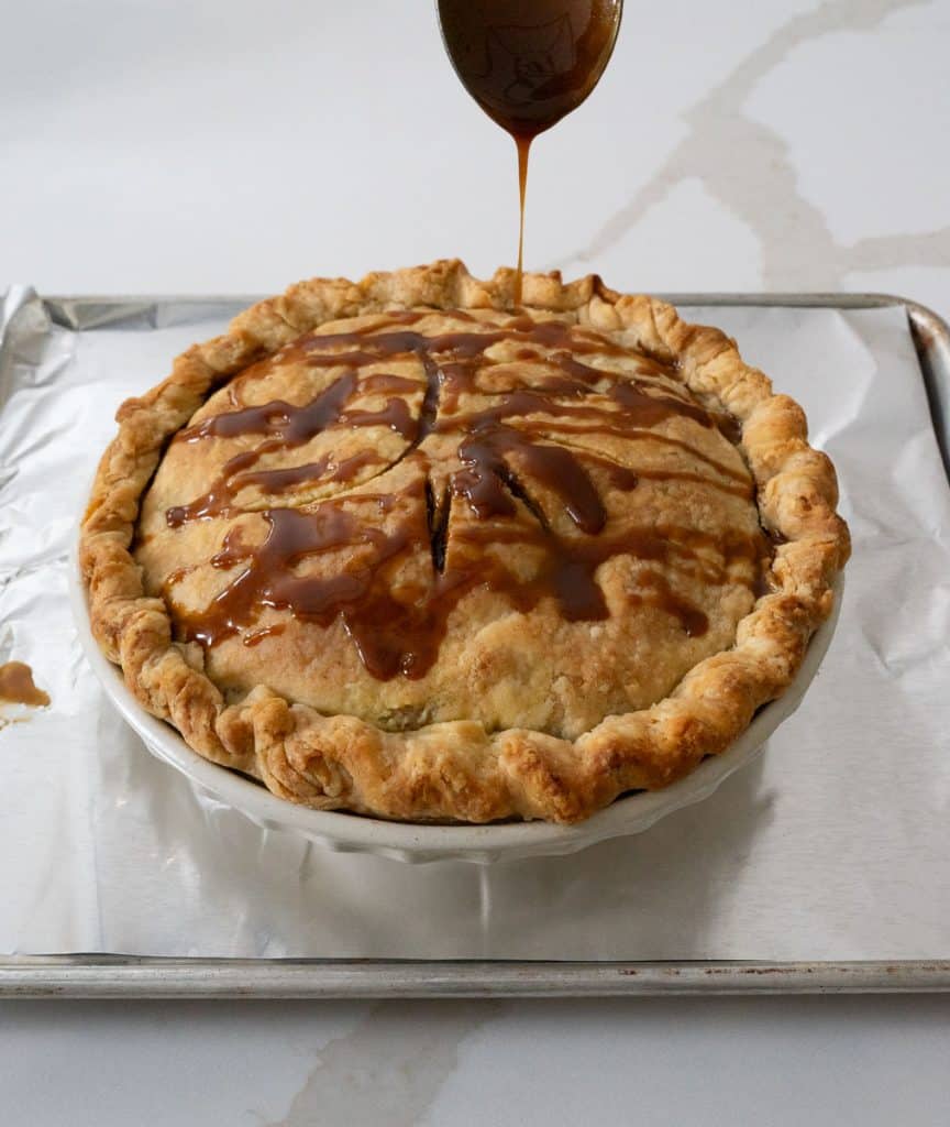 drizzling caramel sauce over top of baked apple pie that is sitting on a baking sheet