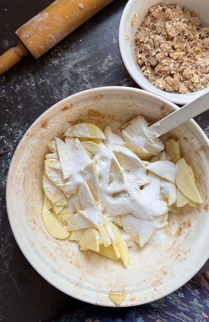 apples in a bowl with sugar and spices on top of them also a bowl of crisp crumble is in the top right of the photo and a rolling pin is in the top left of the photo