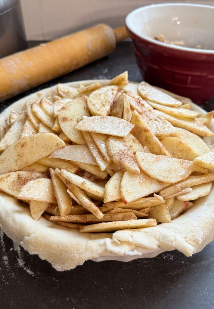 pie crust filled with sliced apples on a black countertop with a rolling pin and a red bowl in the background