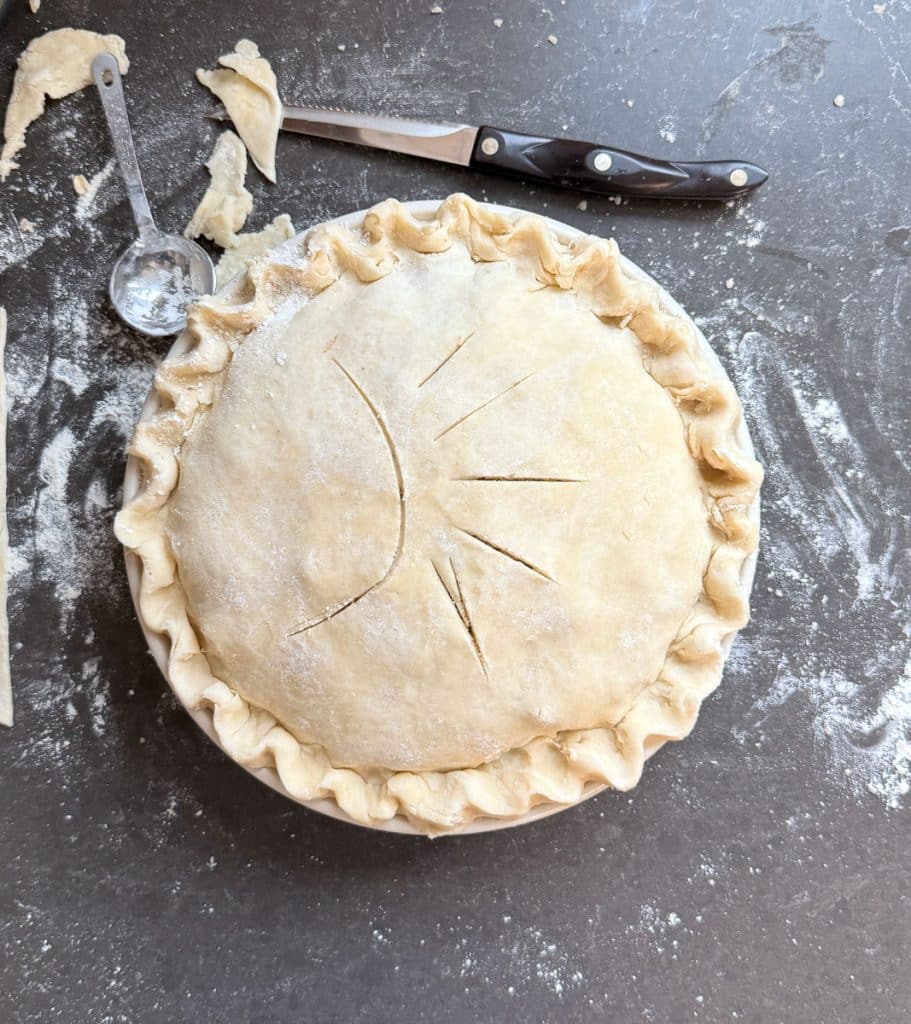 caramel apple crunch pie just before being baked sitting on black countertop with a knife laying in the top of photo with excess pie dough layer around