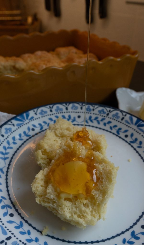 biscuit on a white plate with blue trim. Biscuit has a pad of butter on it and honey is being poured over the biscuit pan of biscuits in background