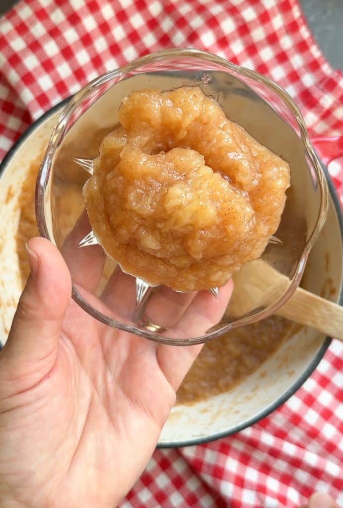 hand holding a glass bowl with applesauce in it over the pot full of apple sauce that is sitting on a red and white gingham cloth