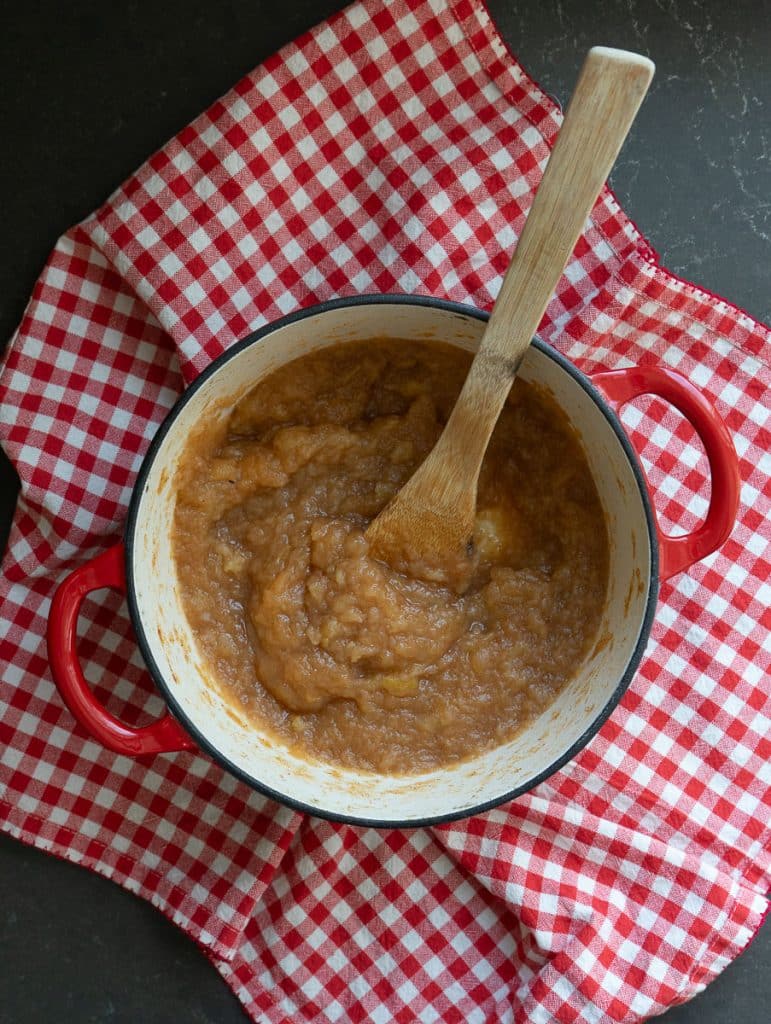 red cast iron pot sitting on a red and white gingham cloth full of applesauce with a wood spoon in the pot of applesauce
