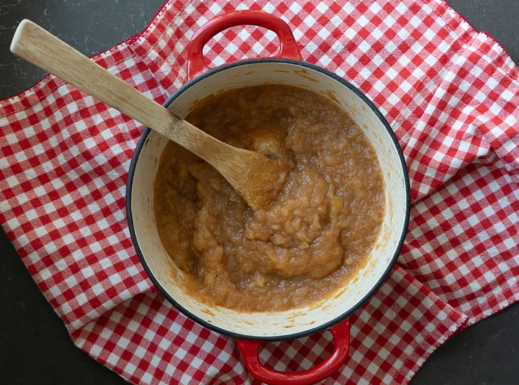 red cast iron pot full of homemade applesauce sitting on a red and white checked table cloth with a wood spoon in the pot