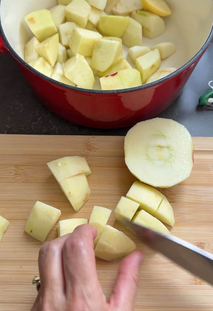 peeling, coring, and cutting apples into chunks, on a wood cutting board the red cast iron pot is at the top of the image with chunks of apples in it