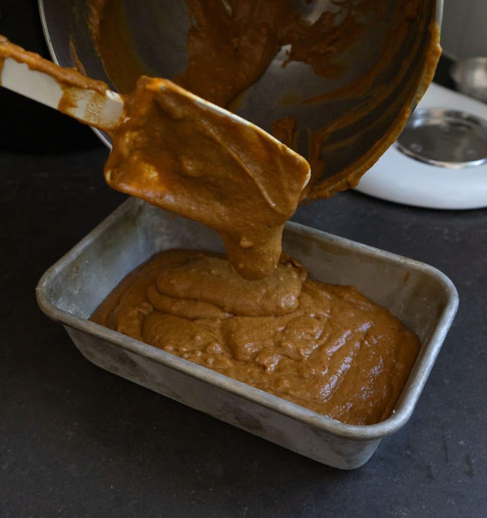 pouring pumpkin gingerbread batter into a loaf pan with spatula scraping the mixing bowl 