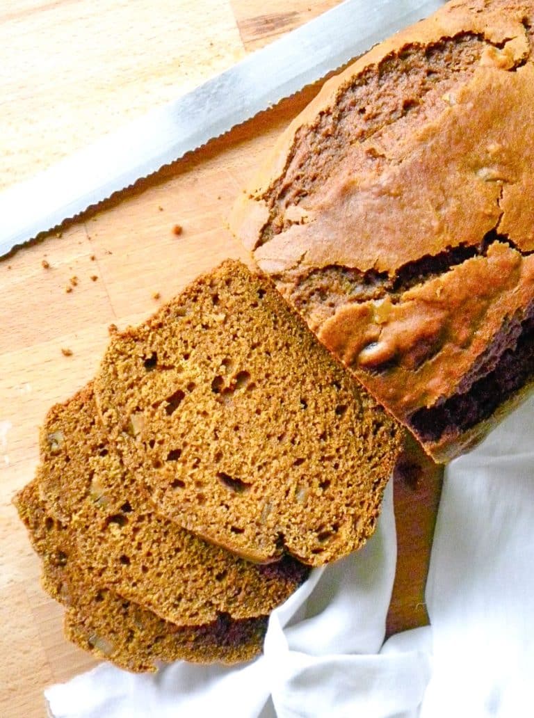 top view of a loaf of pumpkin gingerbread on a wood cutting board sliced with a white tea towel in the lower right corner and a serrated knife in the top of the photo