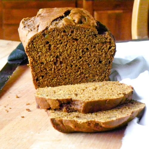 pumpkin gingerbread on a wood cutting board sliced with a white tea towel in the lower right corner and a serrated knife in the top of the photo