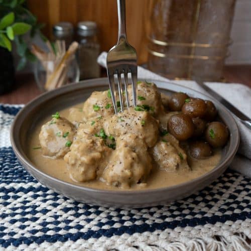 a plate of swedish meatballs on a dinner table with a fork sticking straight into a meatball, served with boiled potatoes