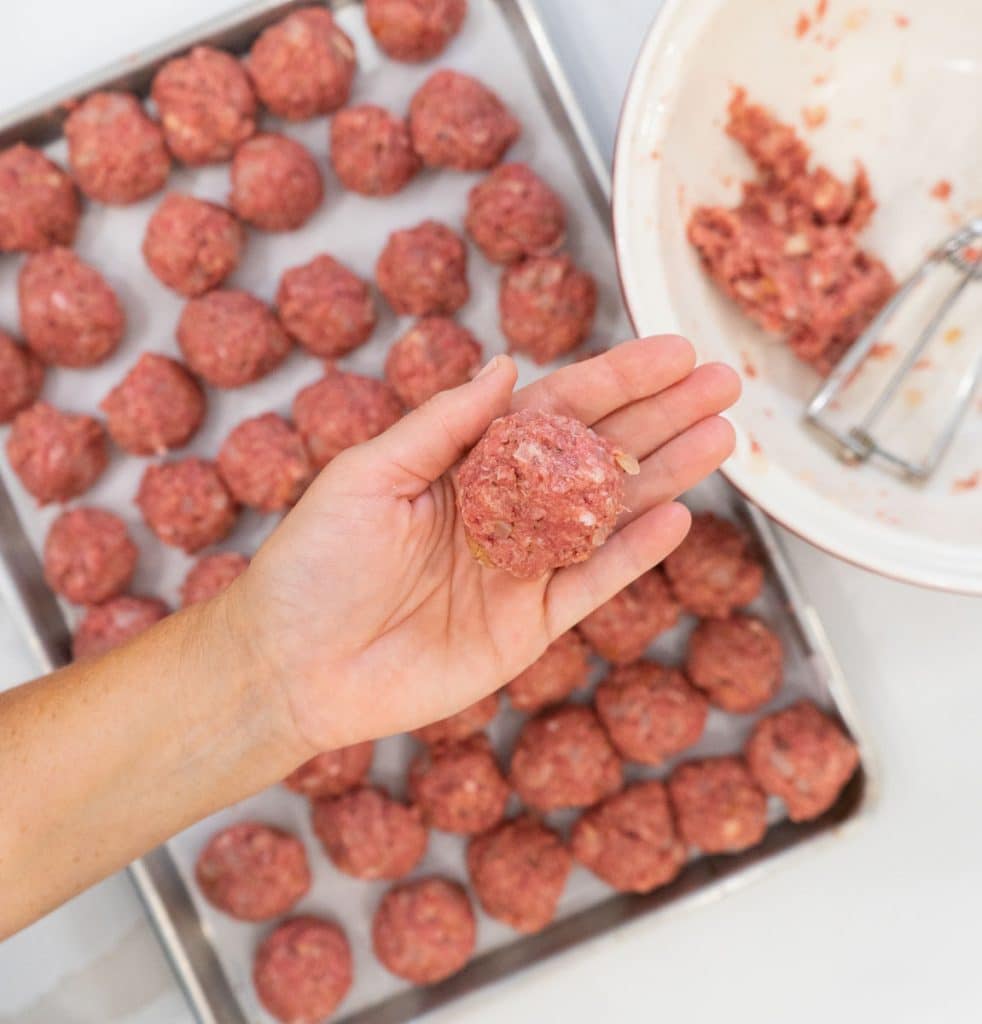 hand holding a raw swedish meatball over a baking sheet of rolled swedish meatballs