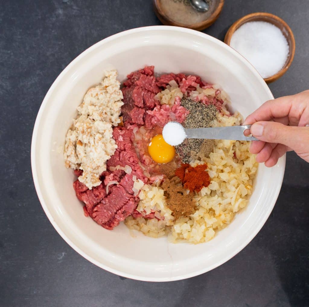 ingredients for swedish meatballs in a large bowl with a hand holding a teaspoon of salt over the bowl.