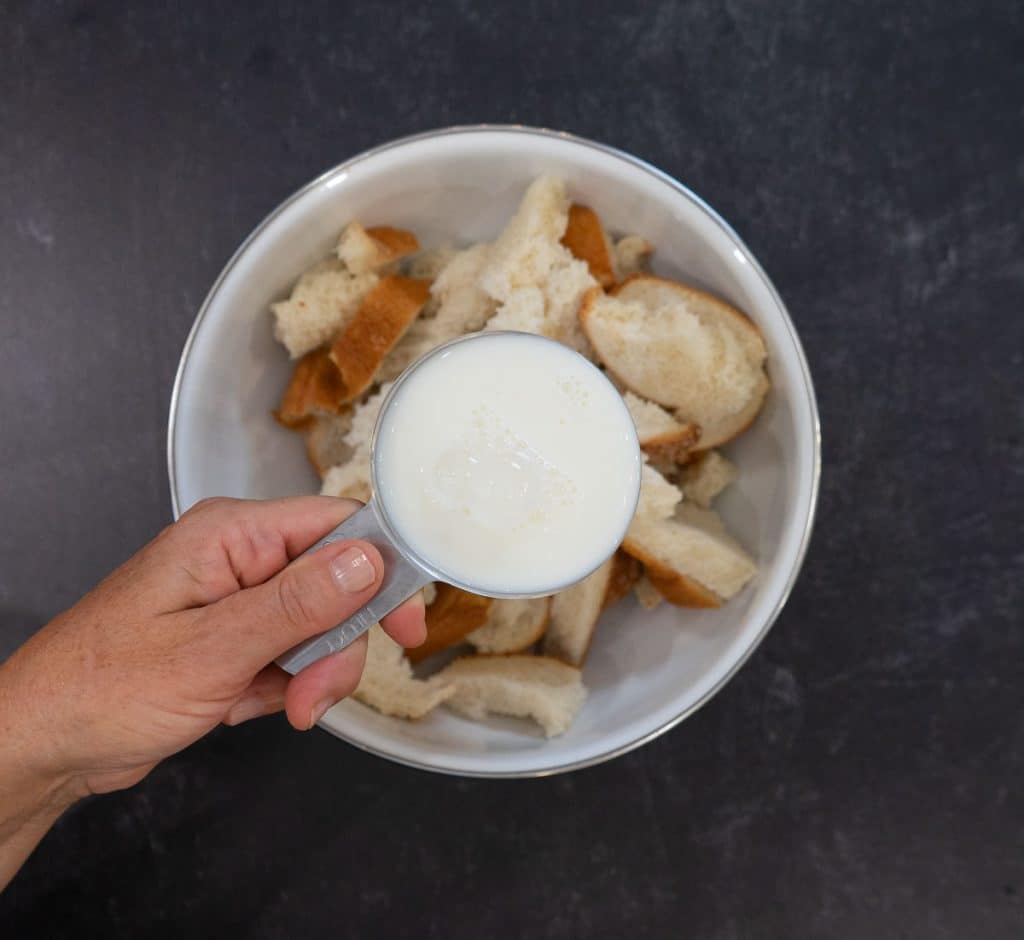 hand holding measuring cup of milk over a bowl of shredded white bread / ingredients for Swedish Meatballs
