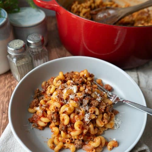 plate of goulash on a tan napkin with the dutch oven of goulash in the back ground salt and pepper shakers are in the upper left corner