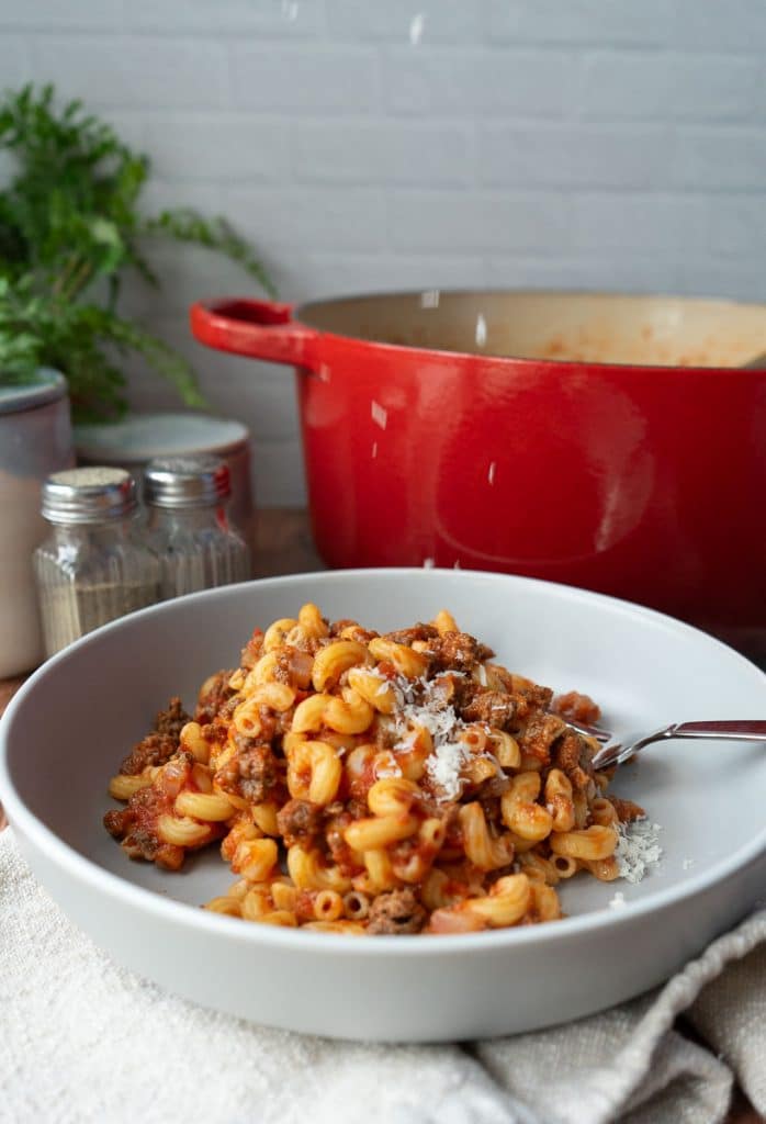 a plate off goulash with a fork on the plate in front of the dutch oven full of goulash salt and pepper shakers are off to the left upper side parmesan is being sprinkled over the top of the pasta
