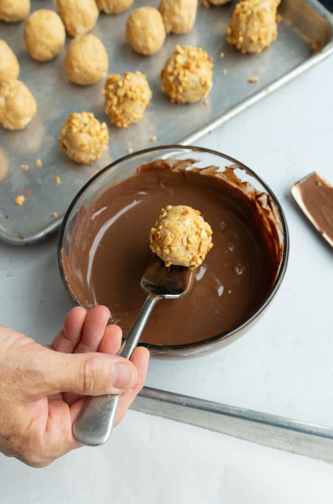 peanut butter ball is on a fork hovering over bowl of melted chocolate pan of peanut butter balls are in the background