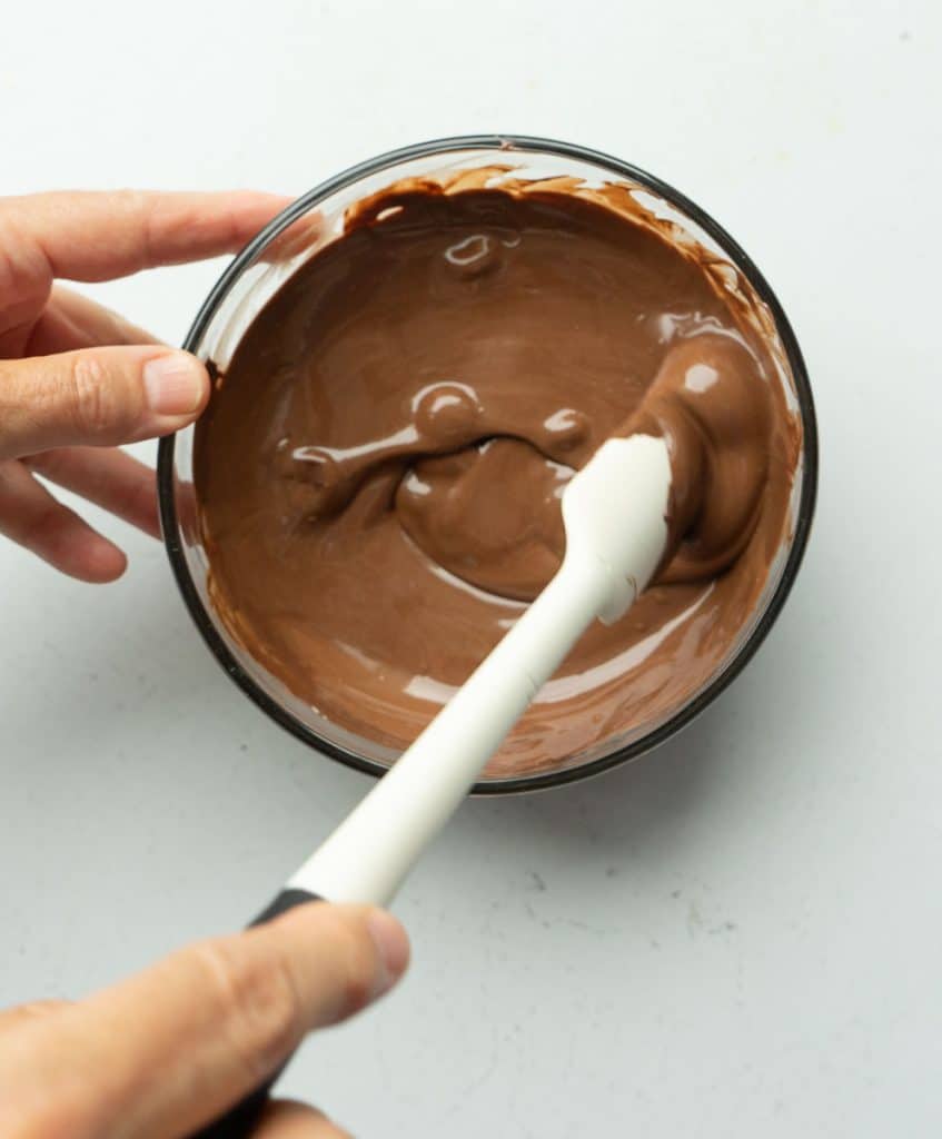 hand on a glass bowl of melted chocolate with a hand stirring chocolate with a white spatula