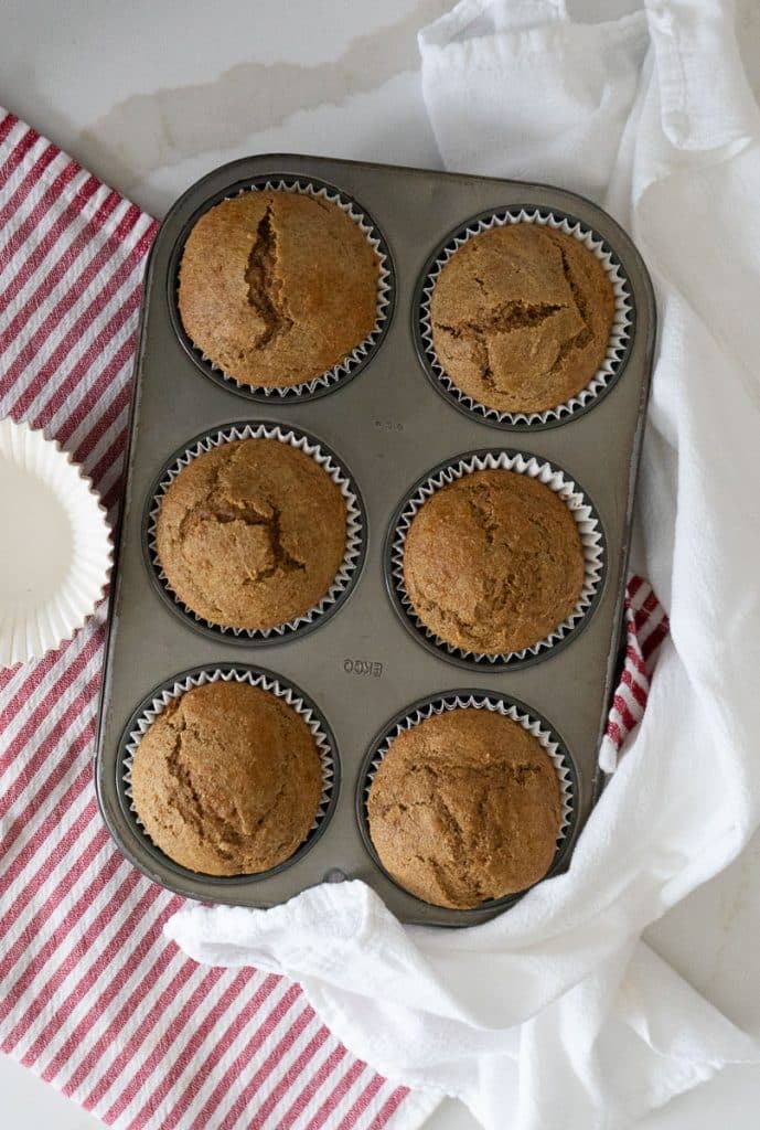baked bran muffin in the muffin tin on a white countertop with muffin papers to the right of the pan and a white and a red and white striped towel under the muffin pan