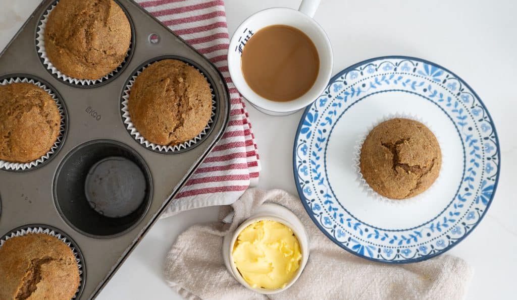 muffin tin with baked muffins in it, a crock of butter, a cup of coffee and then a whole bran muffin is sitting on a white plate with blue flowers around the edge 