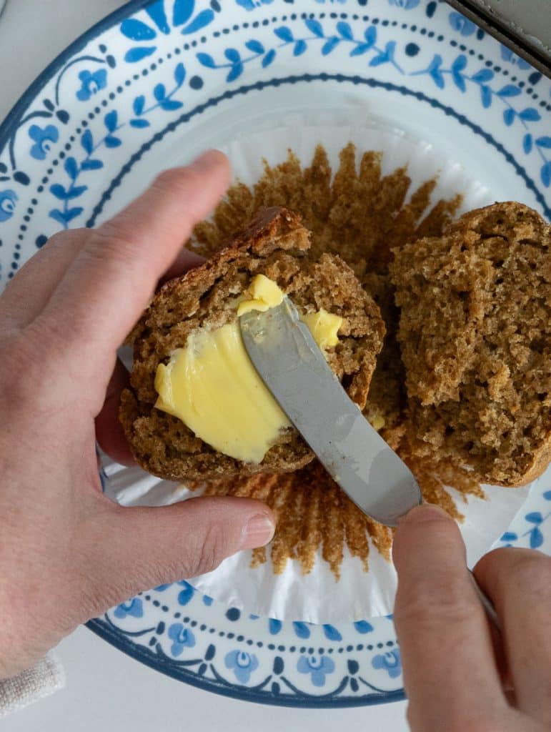 hand holding a half of a bran muffin from the left side of the phone, hand is buttering muffin from the right side, in the back ground is the other half of the muffin on a  white plate rimmed with blue flowers