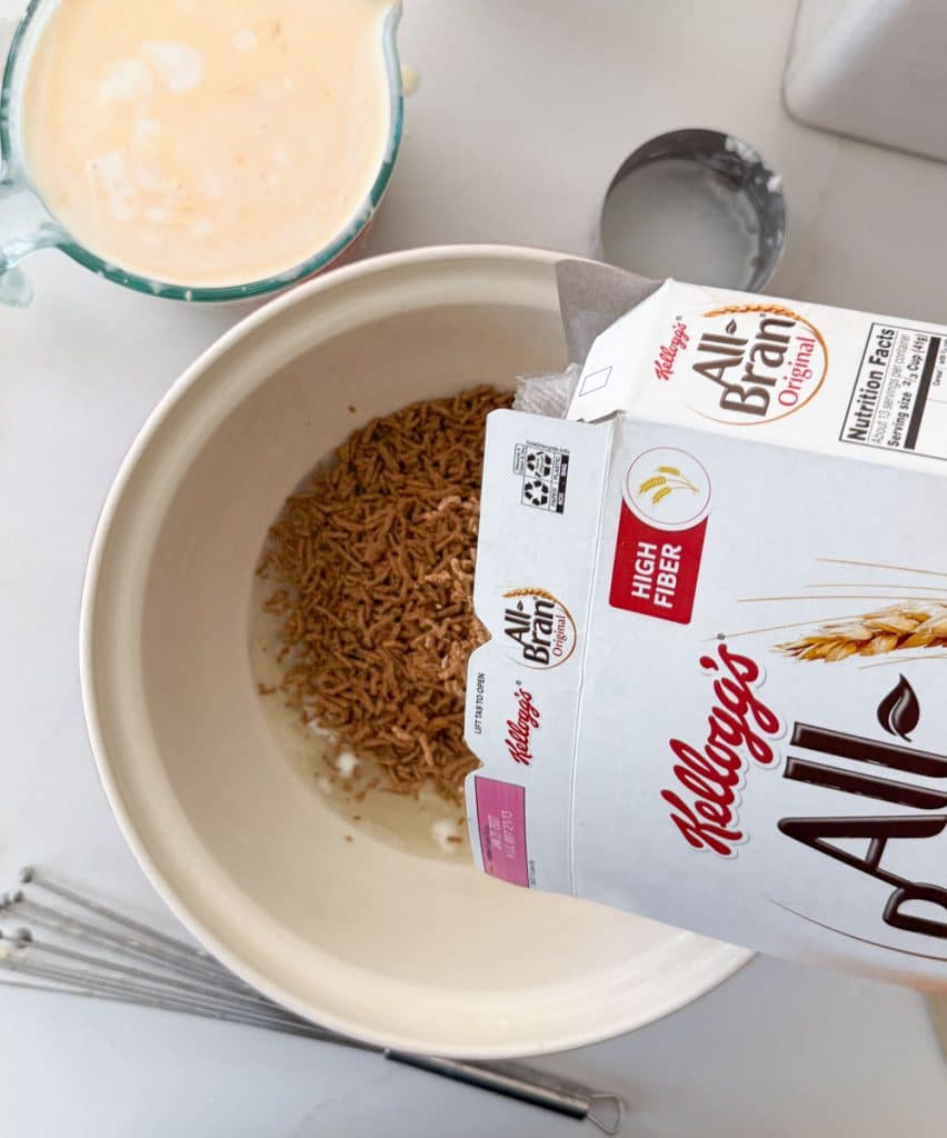 measuring cup of buttermilk is in upper left hand corner, in the middle is a large bowl with a box of all bran cereal being poured into it