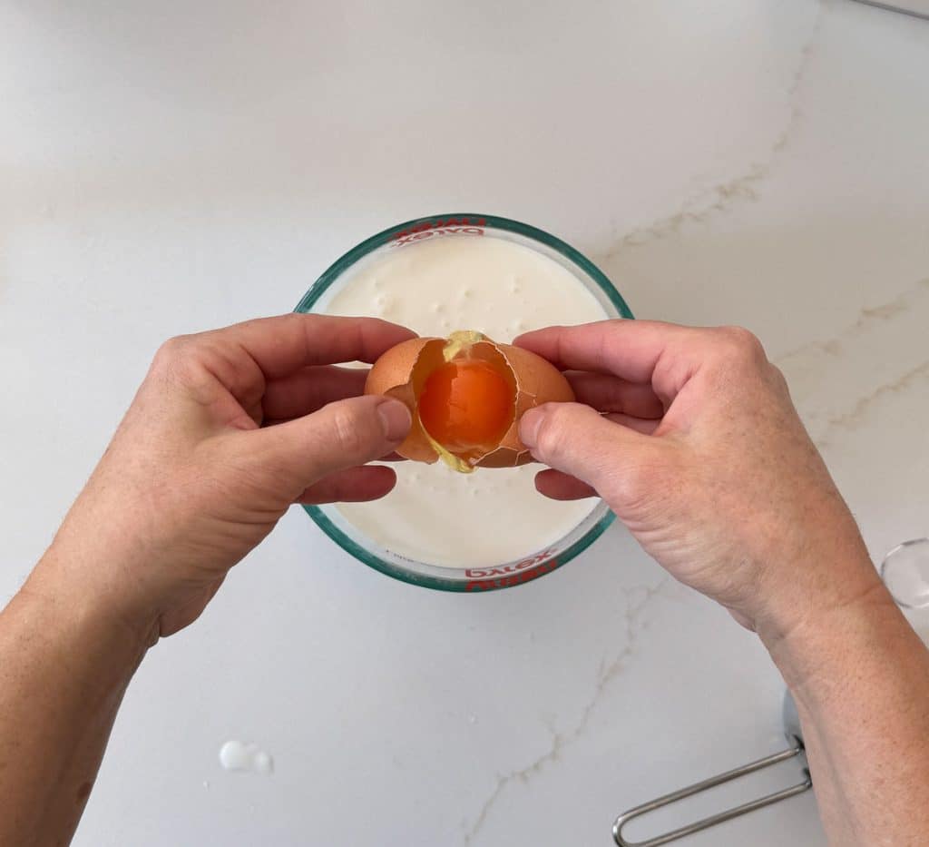 cracking an egg into the buttermilk, photo shows two hands cracking egg over a glass dish of buttermilk on a white countertop