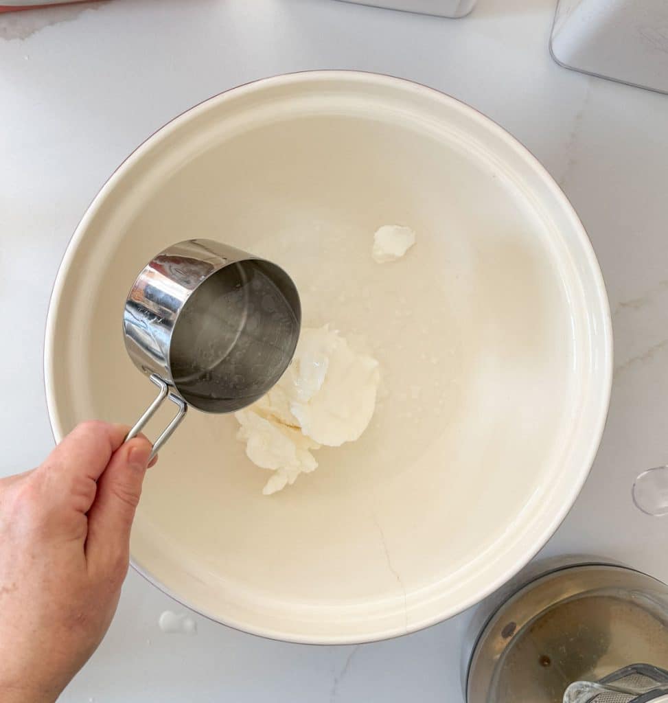 vegetable shortening in a large bowl, with a hand on the left side holding a metal measuring cup with water in it and pouring it into the bowl