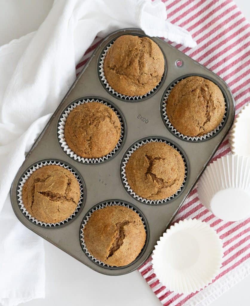 baked bran muffin in the muffin tin on a white countertop with muffin papers to the right of the pan and a white and a  red and white striped towel under the muffin pan