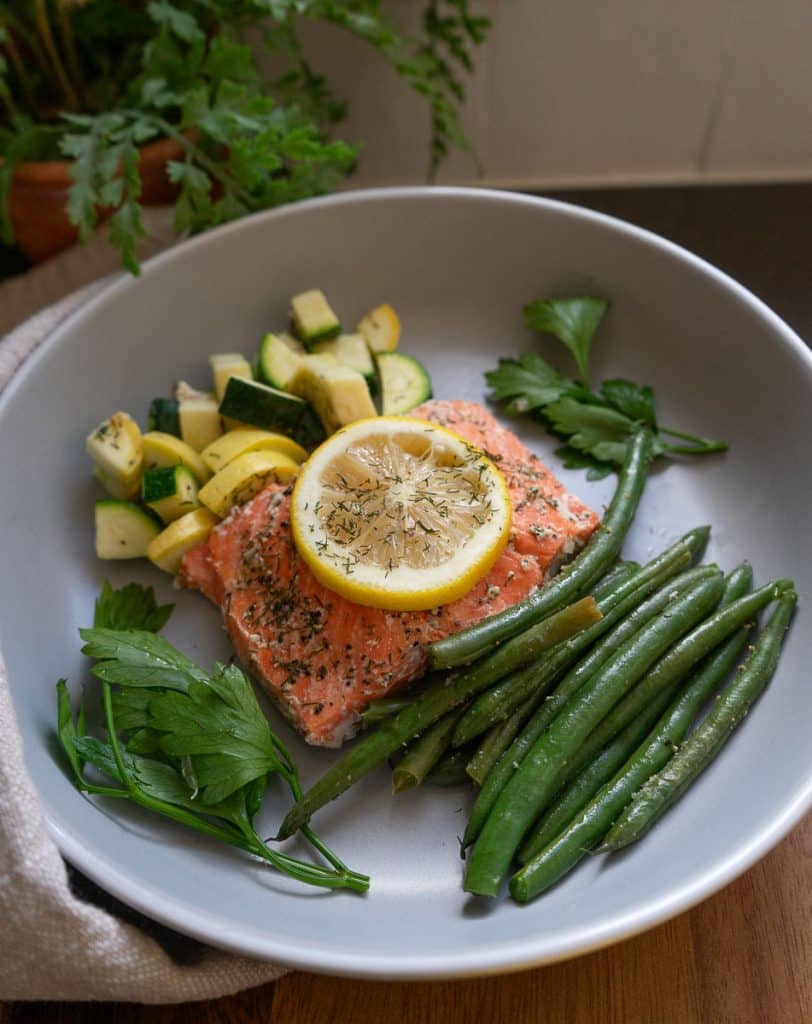 piece of fish / salmon on a grey plate with green beans and zuchini and summer squash. Lemon is on top of the fish. background there are salt and pepper shakers and a plant